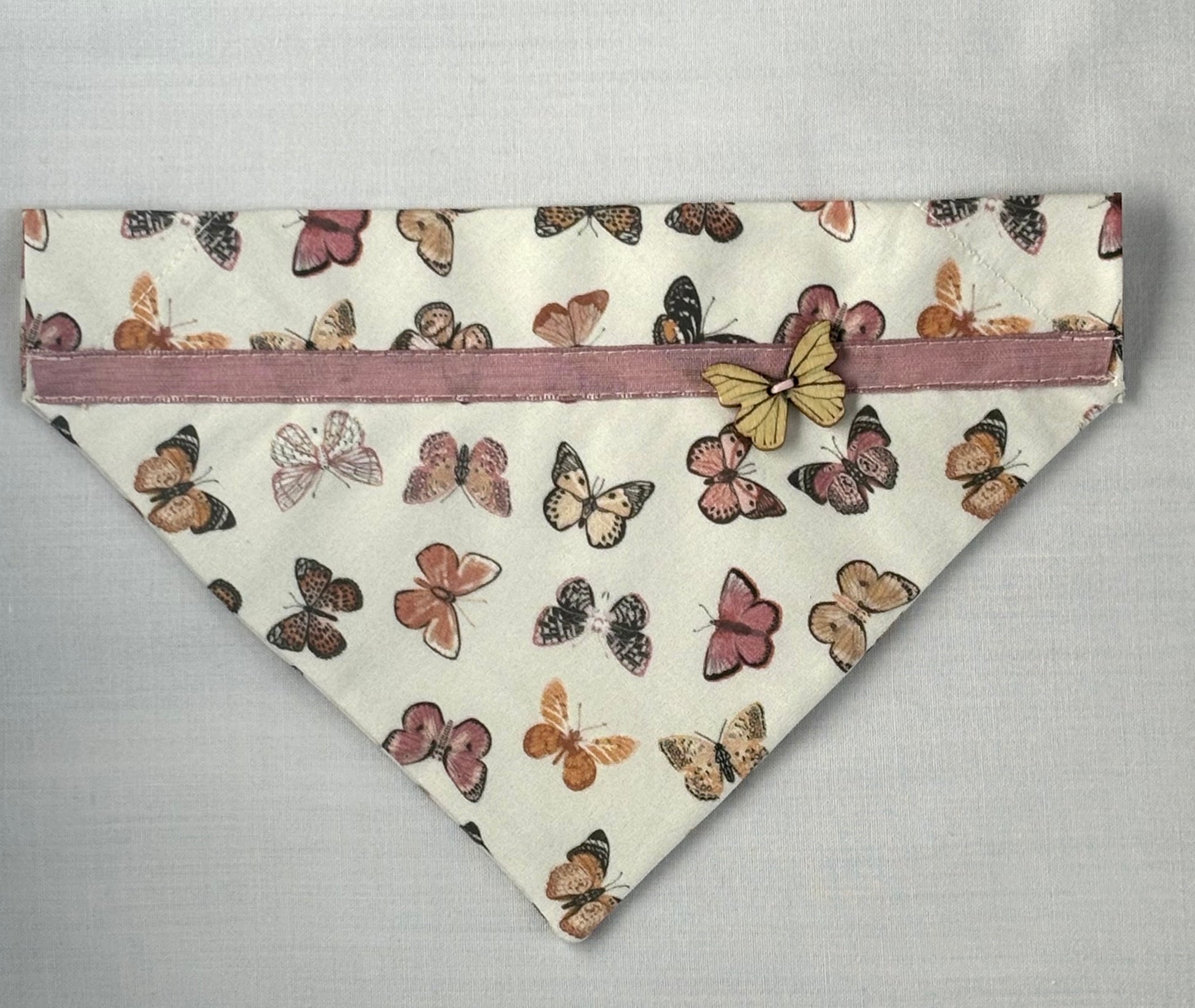 Triangular shaped pet scarf laying flat on a white surface. Scarf has a butterfly print, pink ribbon, and a wooden butterfly button.