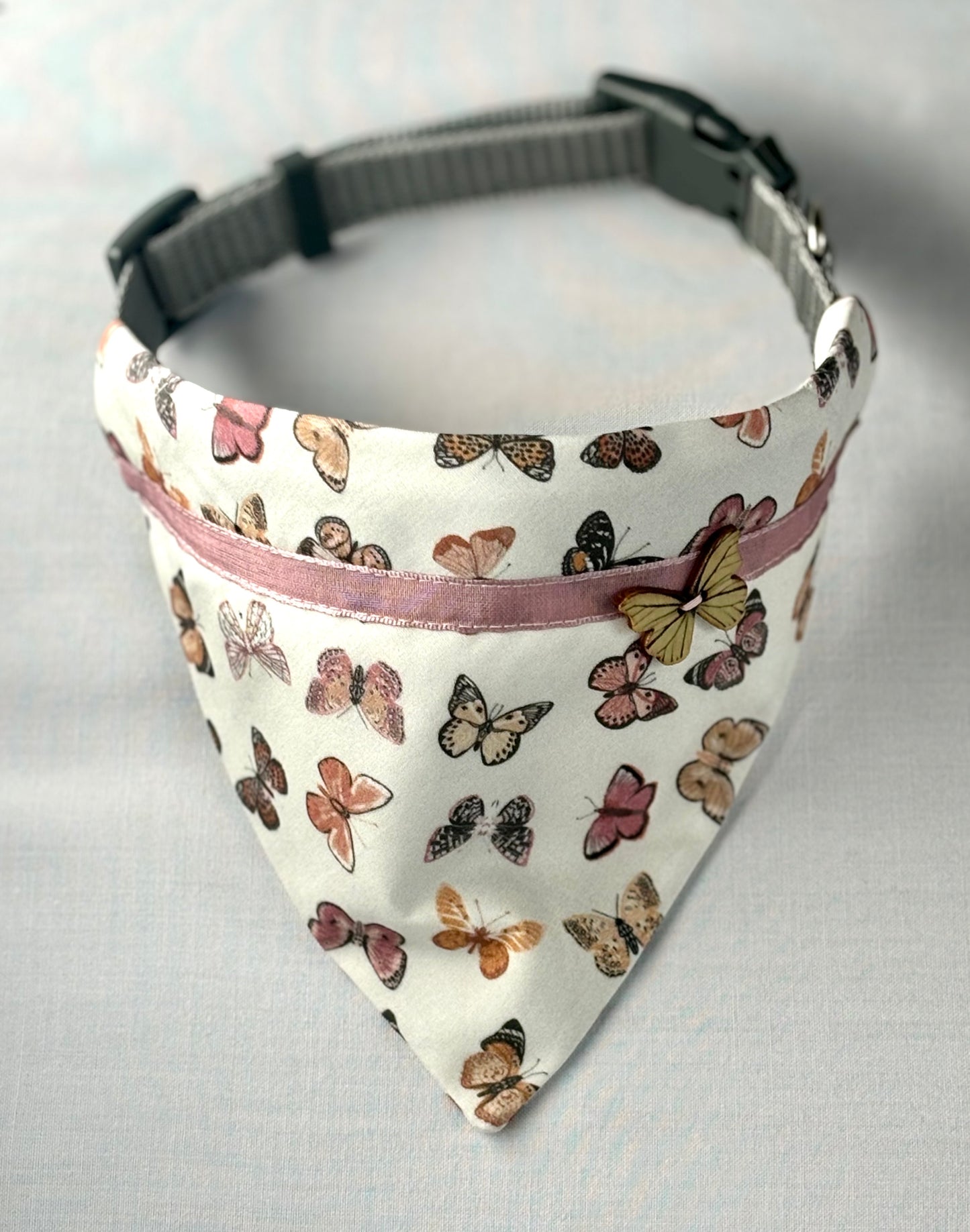 Pet scarf draped over a dog collar sits on a white background. Scarf has a butterfly print, pink ribbon, a butterfly button.