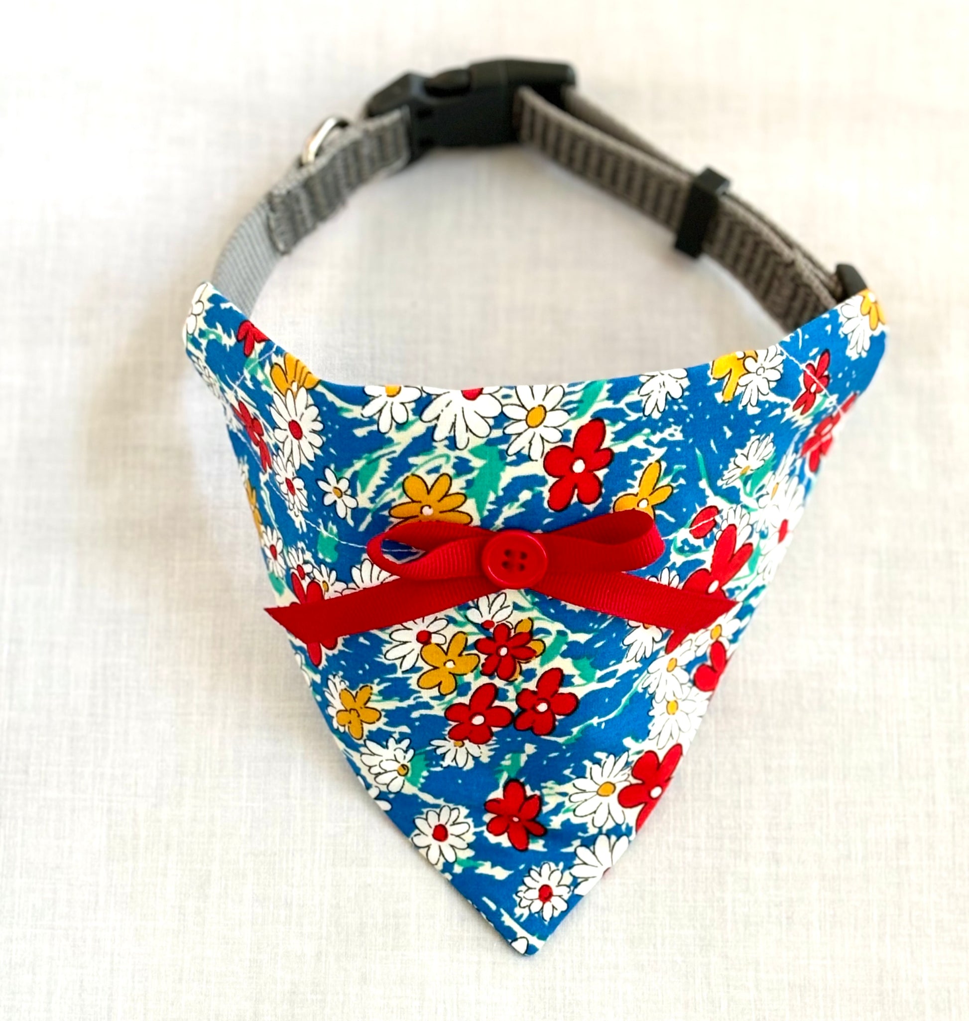 Pet scarf draped over a dog collar sits on a white background. The scarf is royal blue with red, white and blue flowers. Scarf also has a tied bow made from red ribbon and a button sewn at the center of the bow.