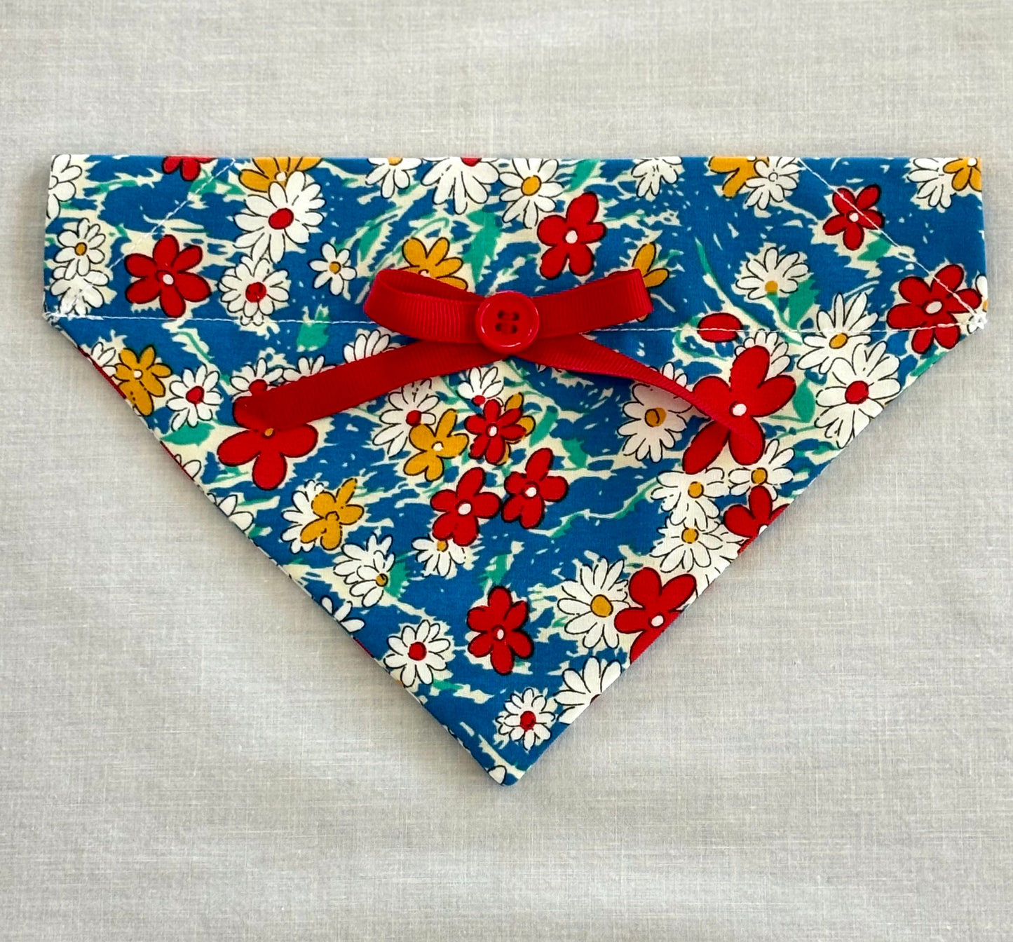 Triangular pet scarf laying flat on a white background.   The scarf is royal blue with red, white and blue flowers. Scarf also has a tied bow made from red ribbon and a button sewn at the center of the bow.